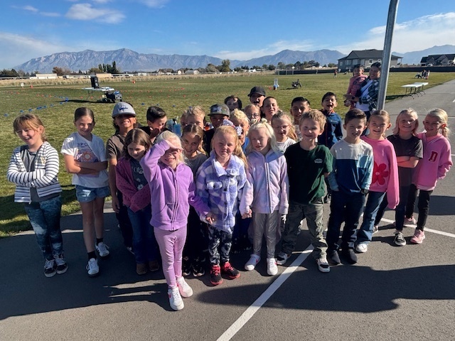 A group of elementary school students are posing together on black asphalt. They are smiling at the camera, with a mountain range and residential area in the background.