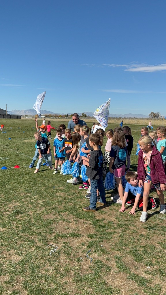 A group of young children and a male adult are gathered on a sunny grass field, possibly at the start of a race. A few children are holding white flags or blue pom-poms. One child's shirt has the word "UTAH" in orange. Mountains are visible in the distance.