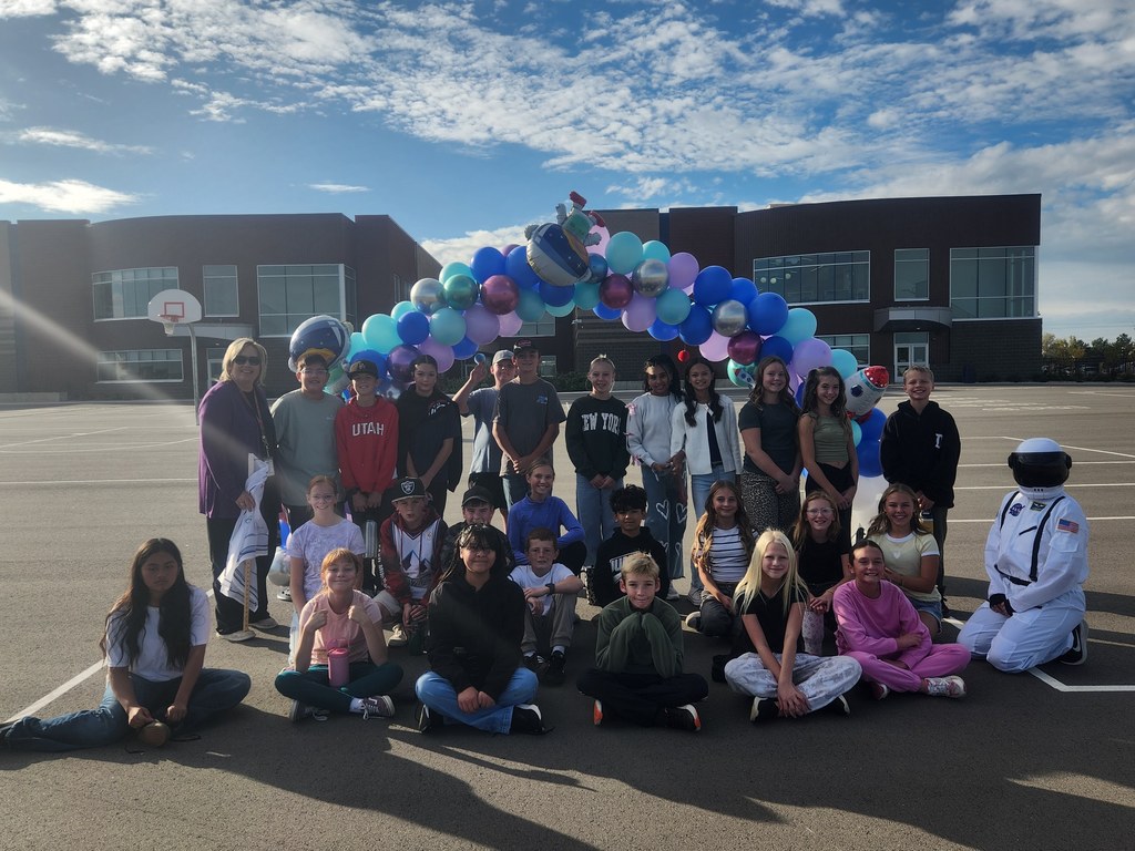 A large group of 6th grade elementary school students are posing for a photo on an asphalt playground in front of a brick school building, underneath a large arch made of blue, purple, and silver balloons. A person dressed in a white astronaut suit is kneeling on the right, and one student's shirt says "UTAH."