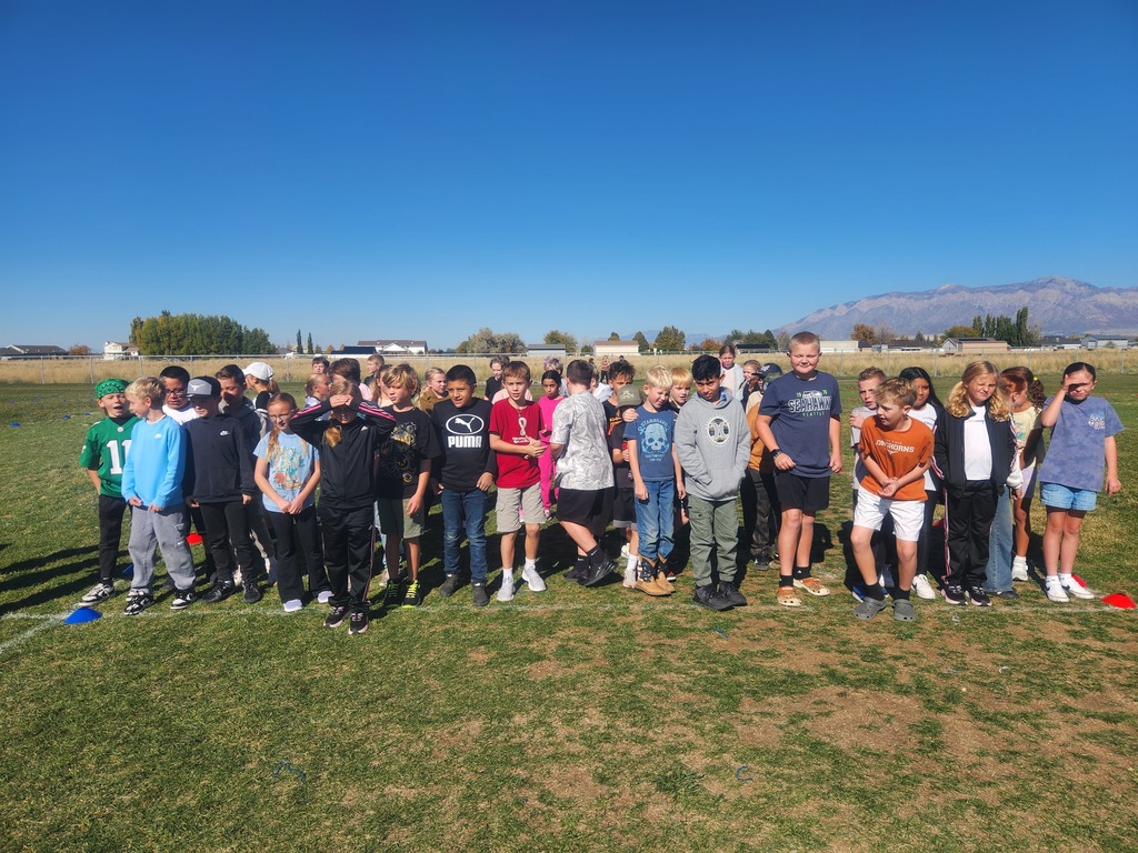 A large line of elementary-aged students are standing still on a grassy field in front of a backdrop of houses and mountains on a bright, sunny day. Some children wear shirts with text like "PUMA" and a brown shirt that says "TEXAS."