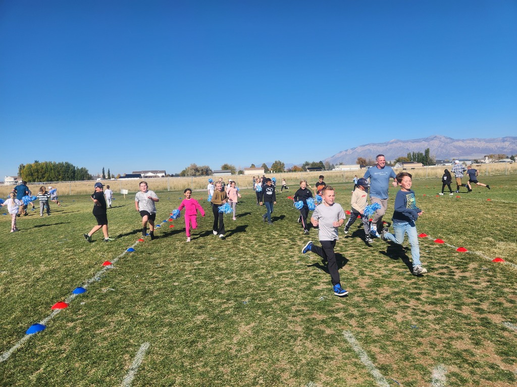 A group of elementary school children and a male adult are running across a grassy field under a clear blue sky. Some children are holding blue pom-poms. Cones mark the edges of the field, and mountains are visible in the background.
