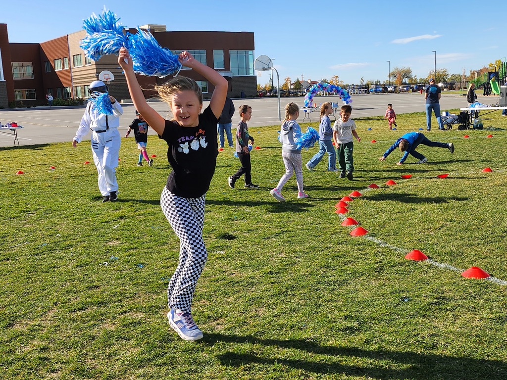 A smiling young girl in a black t-shirt that says "TEACH" with small white images and checkered black and white pants is jumping for joy while holding blue pom-poms high in the air on a sunny day. Other children, a person in a white astronaut-like suit, and a building are in the background on a grassy field with orange cones.