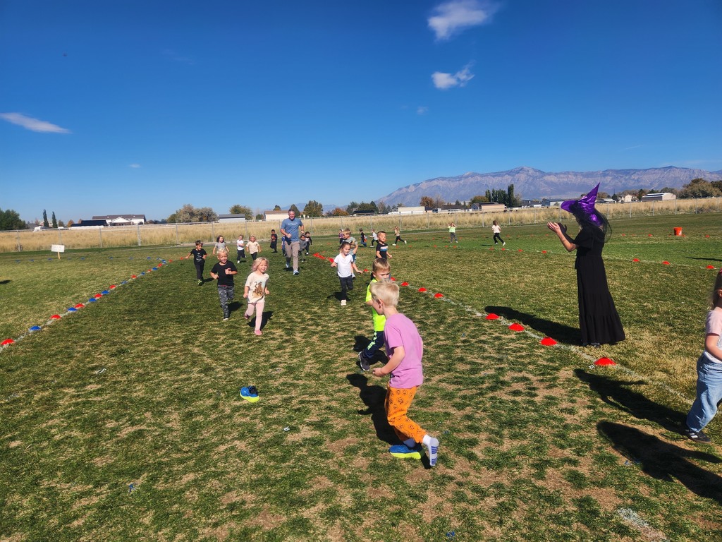Children are running across a sunny, grassy field in a race. A woman dressed in a long black gown and a purple witch's hat is standing on the right, and a male adult is running with the students in the center background. Mountains are in the distance.