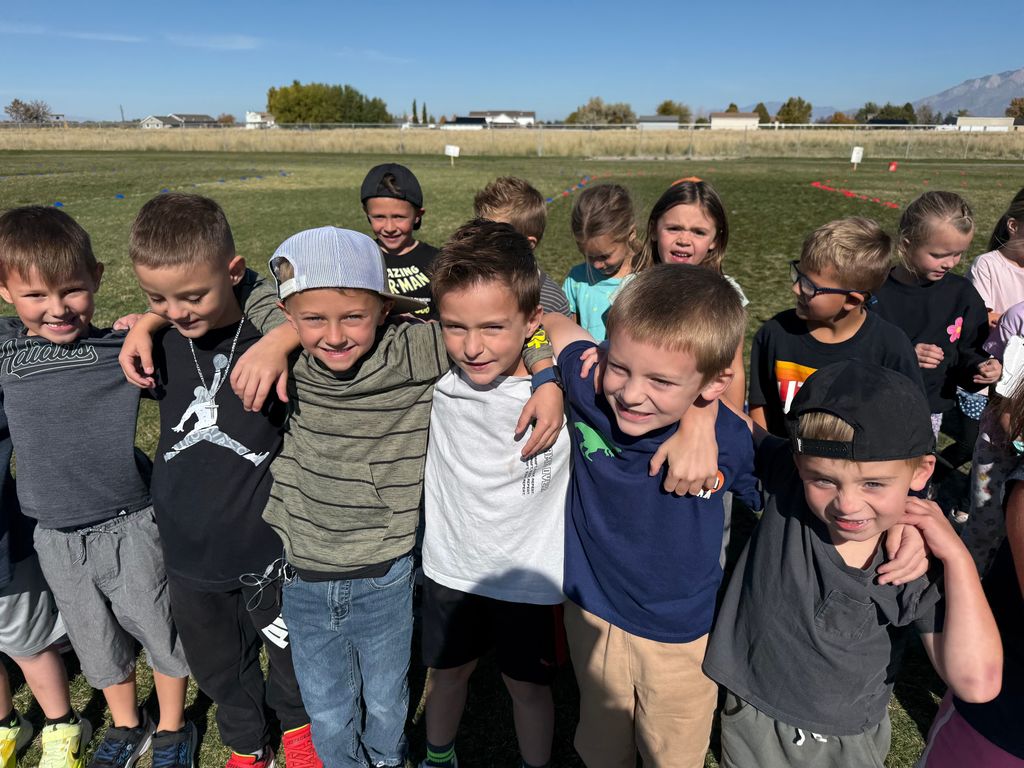A close-up shot of approximately ten young boys smiling and posing for a group photo on a sunny day, with their arms around each other's shoulders. A grassy field and houses are blurred in the background.