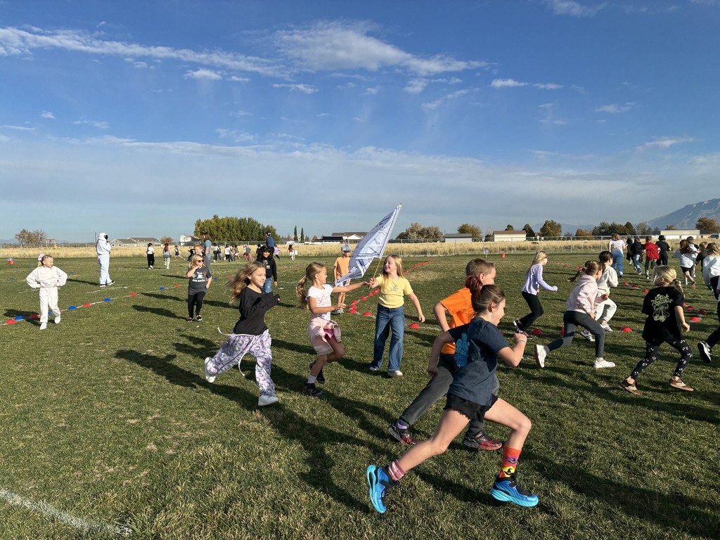 Elementary-aged students are running on a grassy field on a sunny day. In the background, a person in a white astronaut-like suit is walking, and another student is holding a white and blue flag. Mountains are visible in the distance.