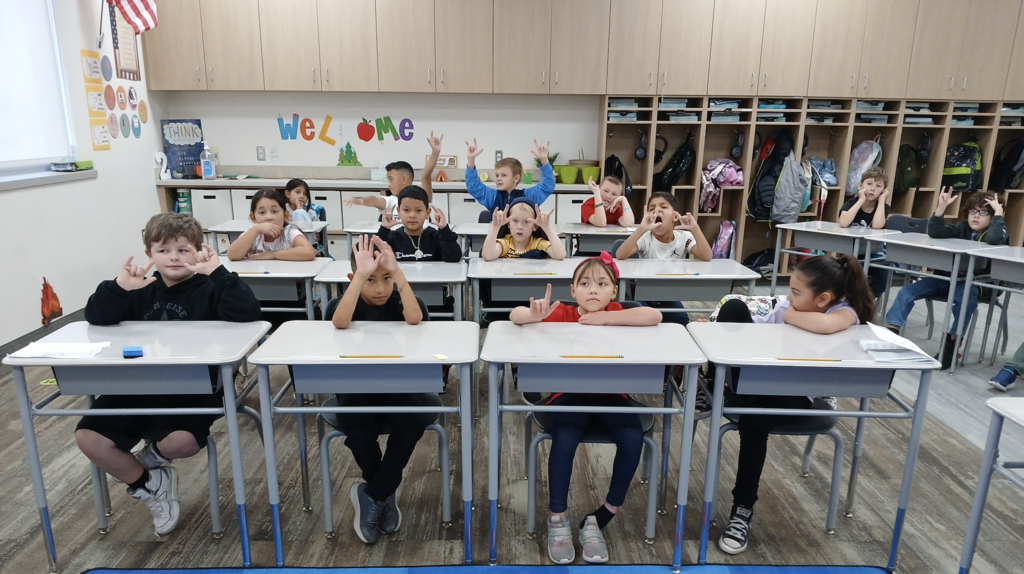 students sitting in desks. Students are showing the I love you symbol