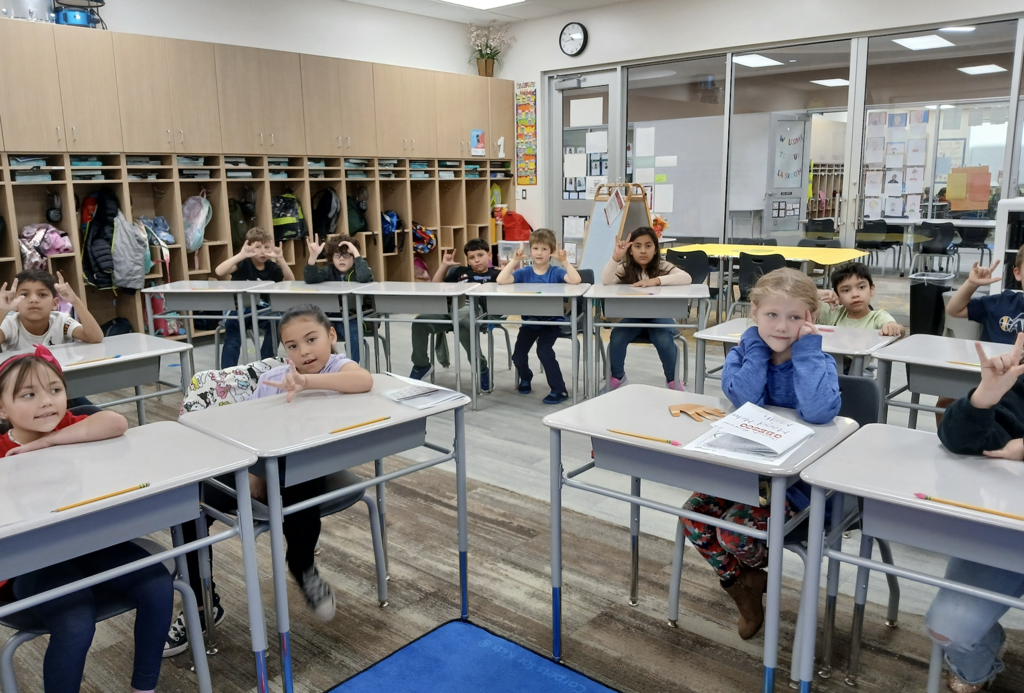 students sitting in desks. Students are showing the I love you symbol
