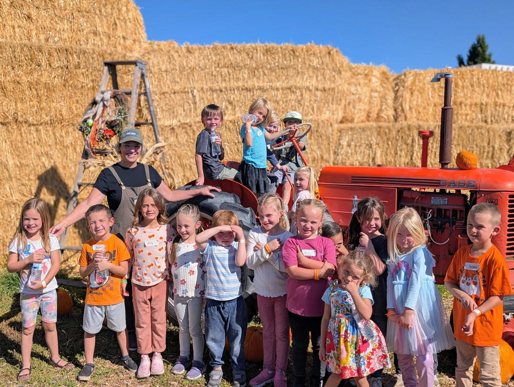 A group of young Lomond View Elementary students and their teacher pose happily for a group photo during a fall field trip. They are standing in front of a large red tractor and a tall wall of hay bales under a clear blue sky.