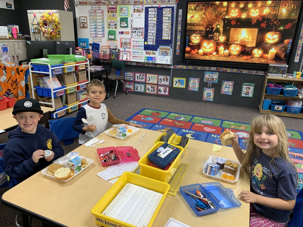 groups of young Lomond View Elementary students smiling and eating lunch together at their desks and tables inside their cozy, fall-decorated classroom.