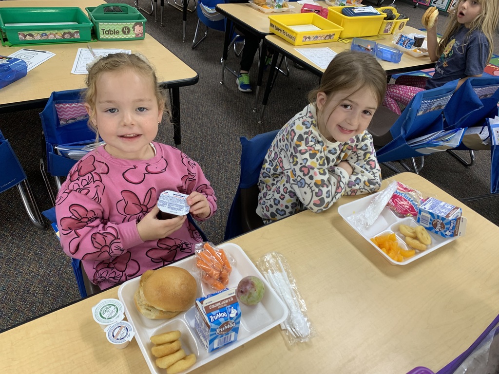 groups of young Lomond View Elementary students smiling and eating lunch together at their desks and tables inside their cozy, fall-decorated classroom.