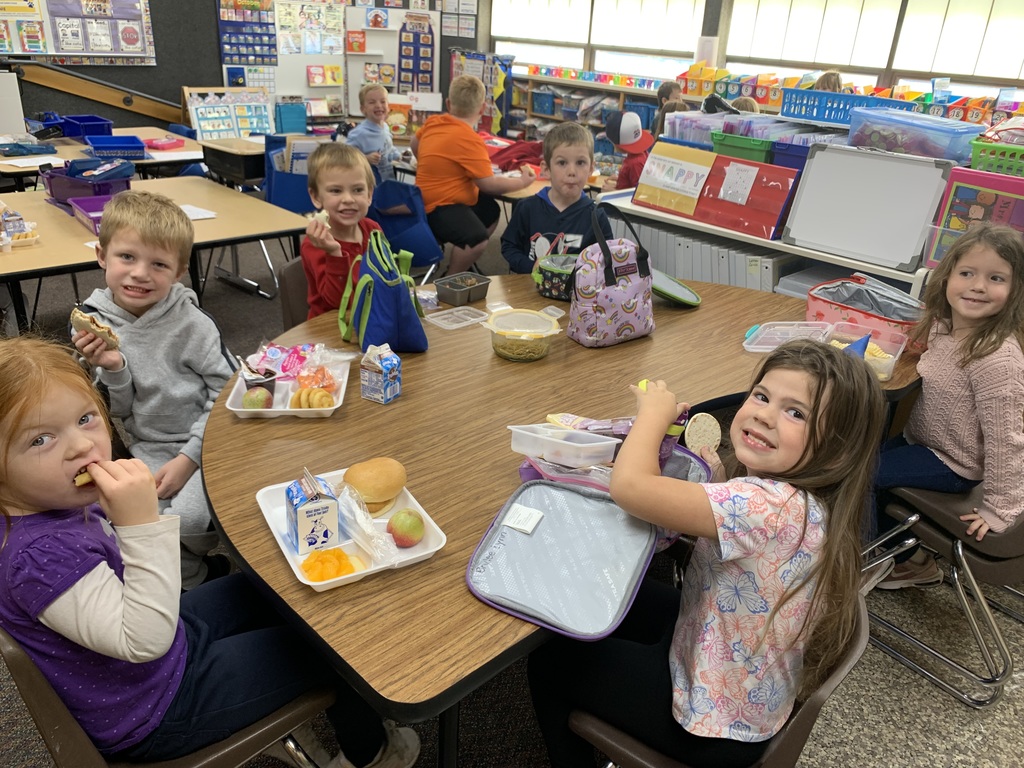 groups of young Lomond View Elementary students smiling and eating lunch together at their desks and tables inside their cozy, fall-decorated classroom.