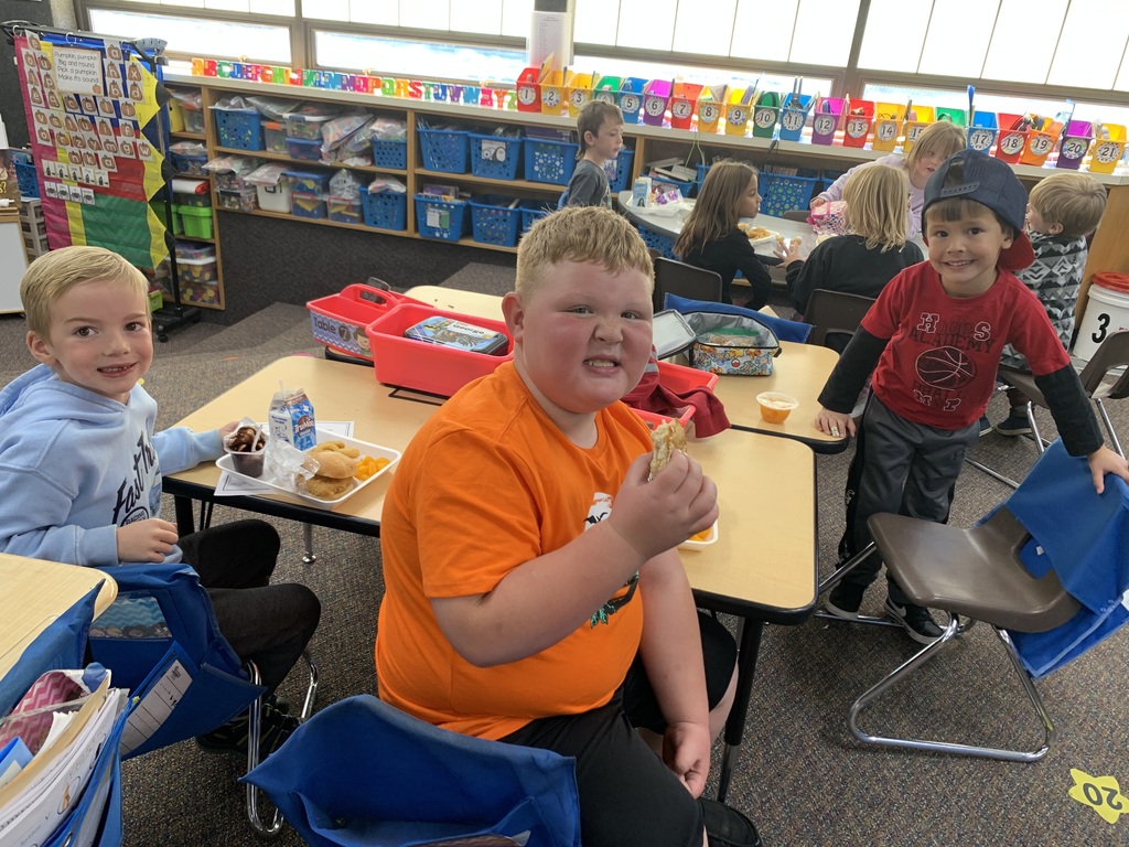 groups of young Lomond View Elementary students smiling and eating lunch together at their desks and tables inside their cozy, fall-decorated classroom.