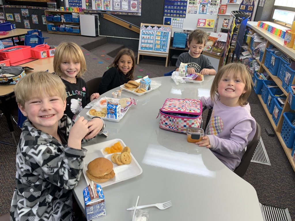 groups of young Lomond View Elementary students smiling and eating lunch together at their desks and tables inside their cozy, fall-decorated classroom.
