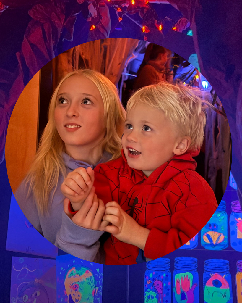 Two children, an older girl and a young boy in a Spider-Man costume, look up in awe inside a dark room filled with glowing neon decorations at a school event.