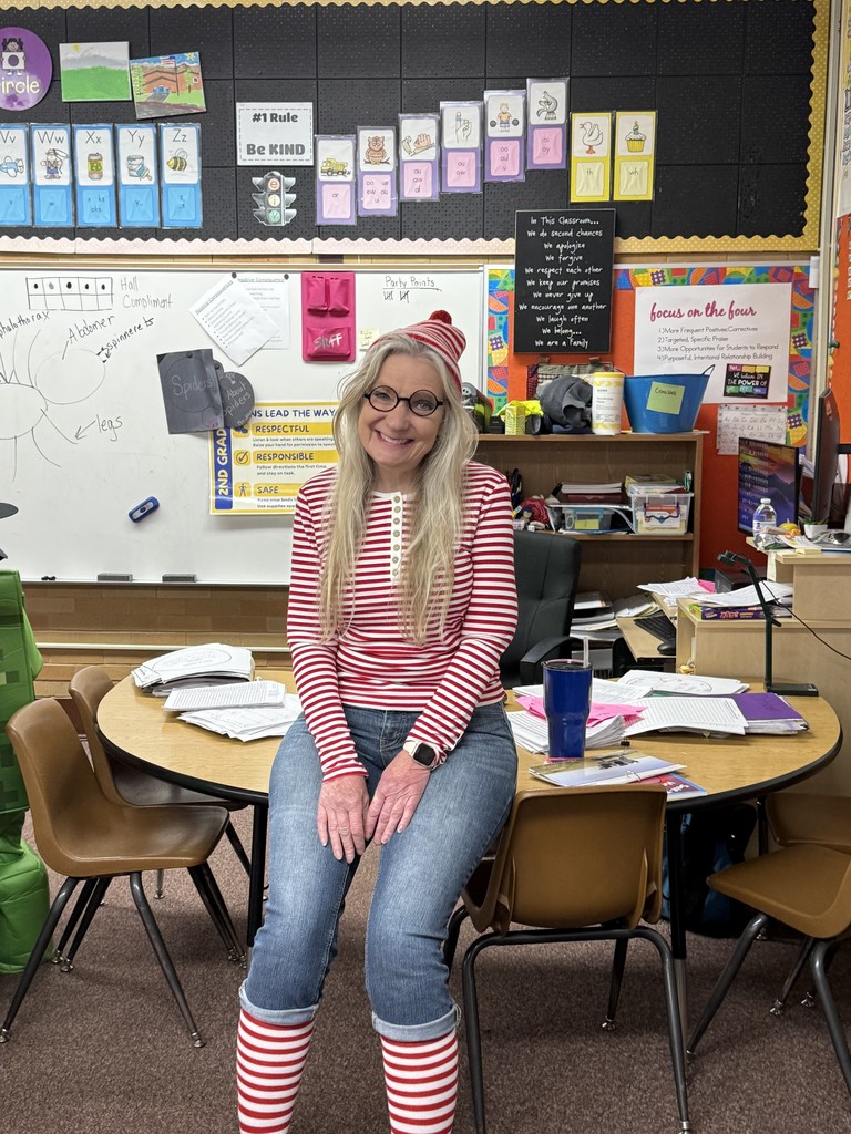 A woman dressed as the character Waldo/Wally (red and white stripes, glasses, hat) standing in a school hallway in front of lockers with a "Who Are We?" sign.