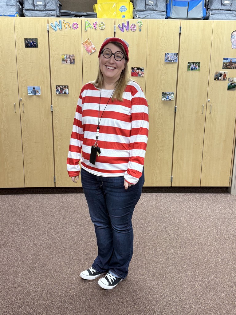 A woman dressed as the character Waldo/Wally, sitting on a round table in a colorful classroom.