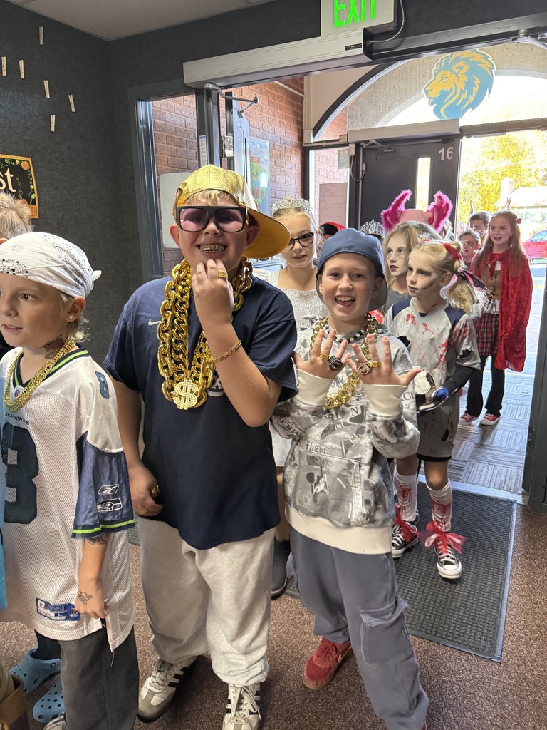 Two elementary school boys in hip-hop/rapper costumes with gold chains, sunglasses, and baseball caps, smiling and posing in a line of students in various costumes.