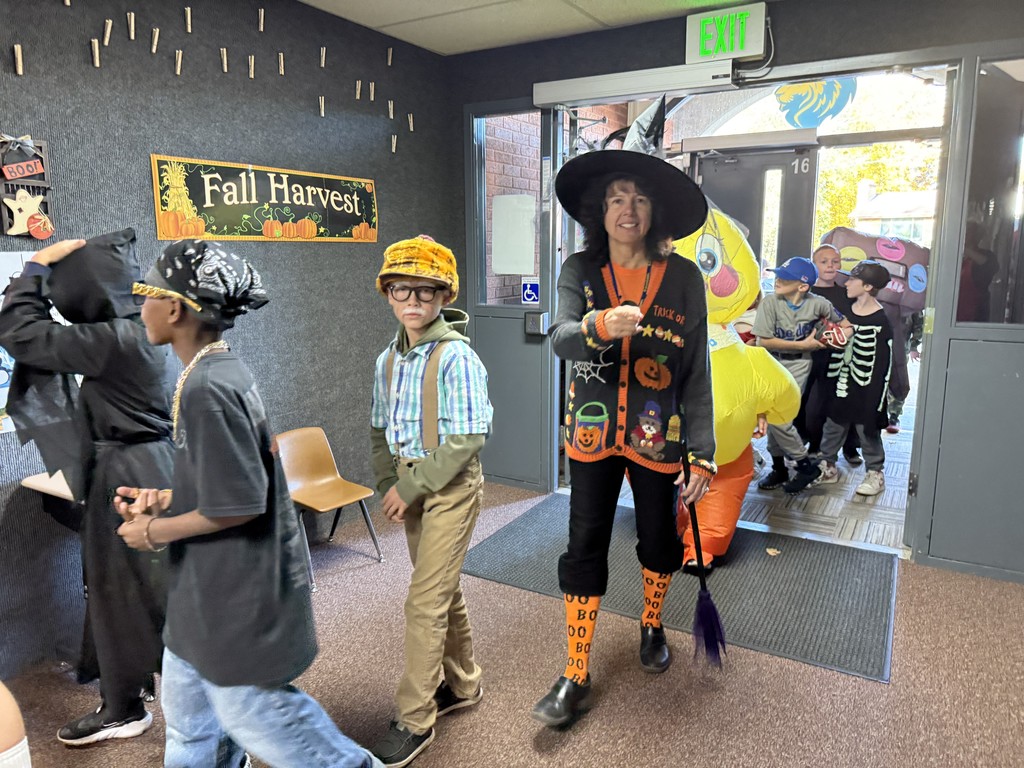 A line of students in costumes (including a ghoul, a scarecrow/farmer, and a large inflatable yellow costume) following a teacher dressed in a Halloween-themed black sweater, witch's hat, and orange socks, holding a small broom.