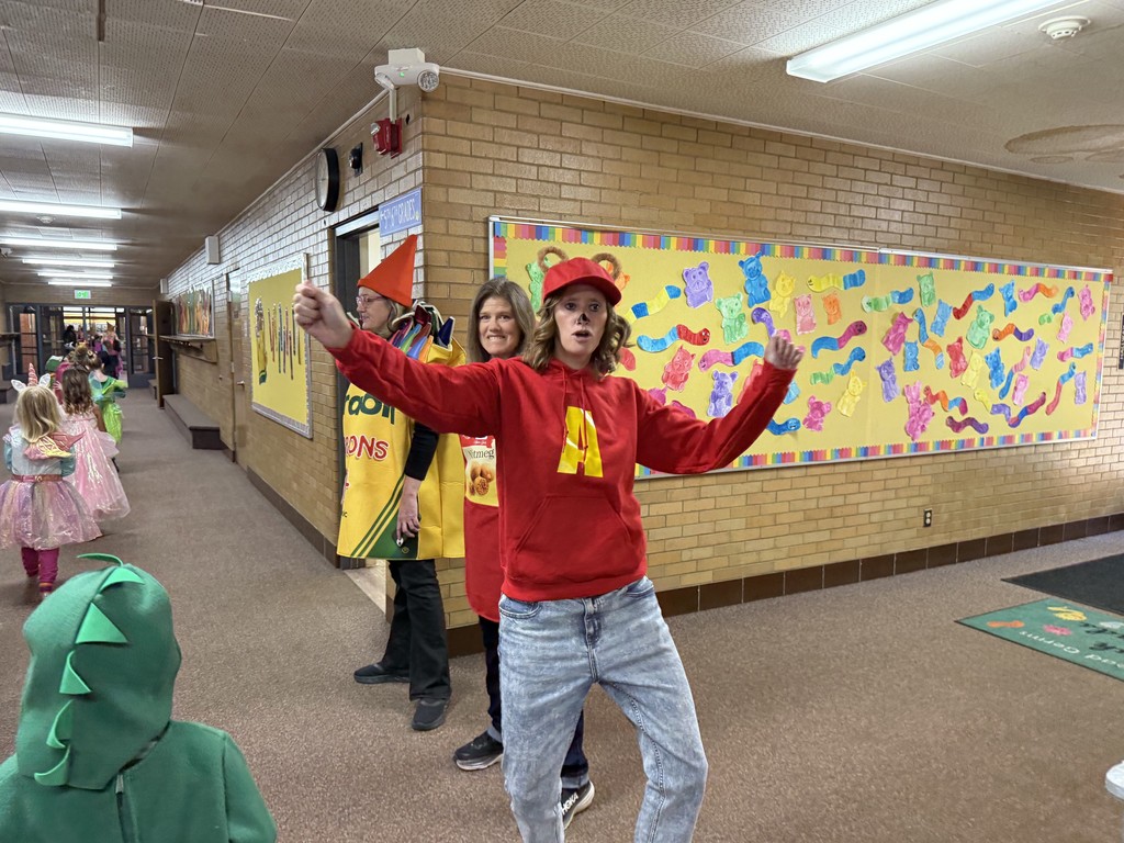 A teacher dressed as Alvin from Alvin and the Chipmunks (red "A" hoodie and baseball cap) posing dynamically in a school hallway. Another teacher in a yellow crayon costume is behind her.
