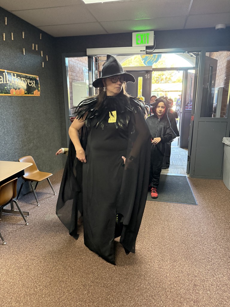 A teacher dressed in a long black feathered cloak and witch's hat, walking through a doorway with a student dressed as a bat behind her.