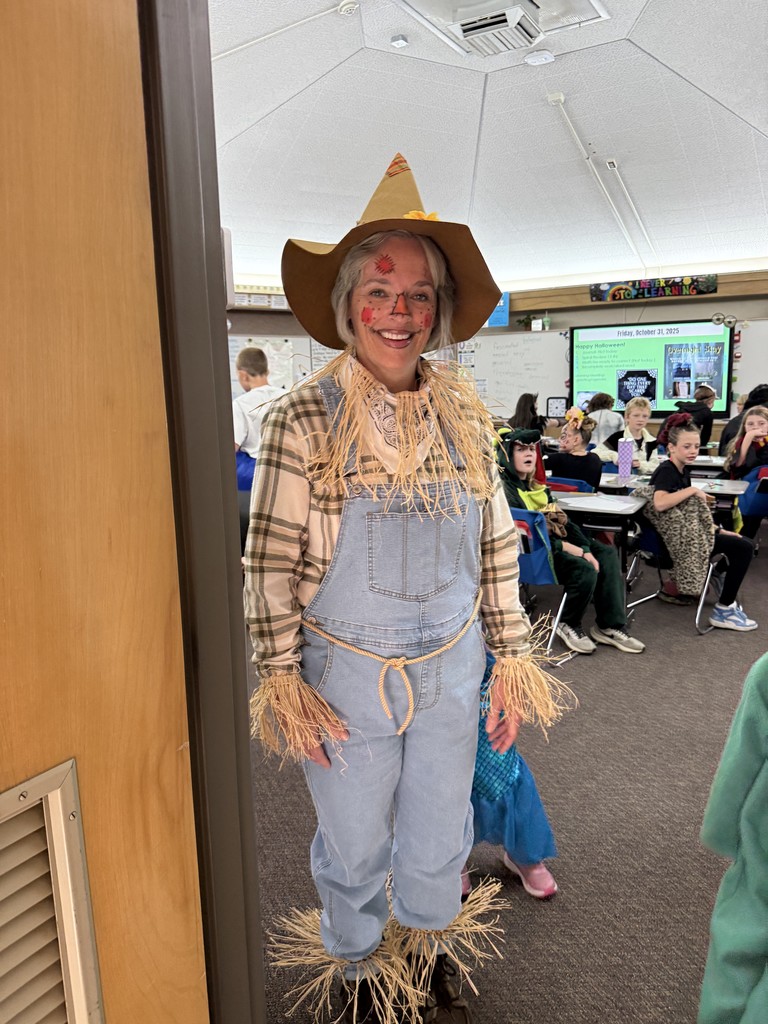 A woman dressed as a scarecrow (overalls, plaid shirt, straw attachments, painted cheeks, and a straw hat) smiling at the camera near a classroom doorway.