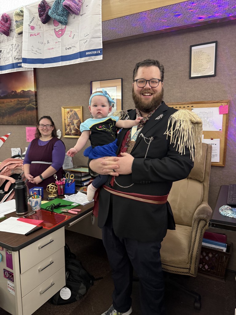 A man in a royal or military-style costume holding a baby girl dressed as Princess Anna from Frozen. A woman in a purple shirt stands smiling in the background.