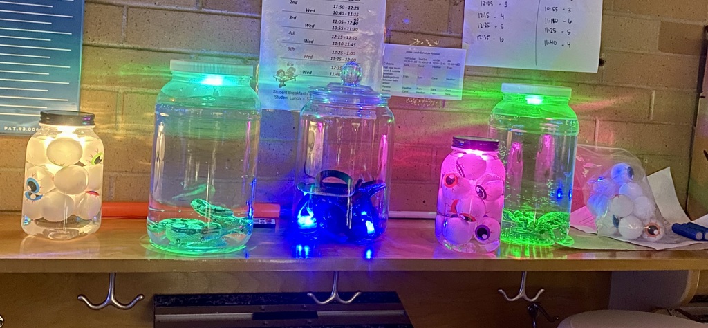 A close-up of several clear jars displayed on a shelf, decorated with eyeballs and snakes and illuminated with bright green, pink, and blue LED lights for a spooky Halloween effect.