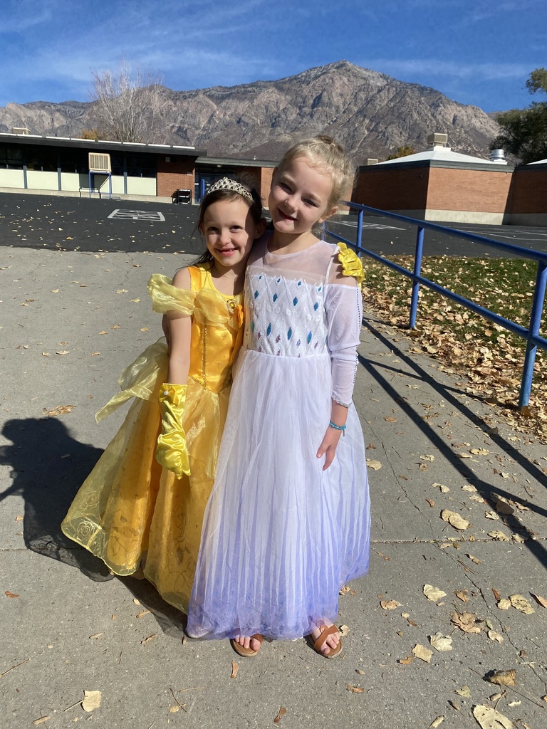 Two young elementary school girls standing outside on a sunny day with a mountain backdrop, hugging and smiling. They are dressed as Disney princesses: one in a yellow Belle gown, and the other in a white and purple Elsa dress.
