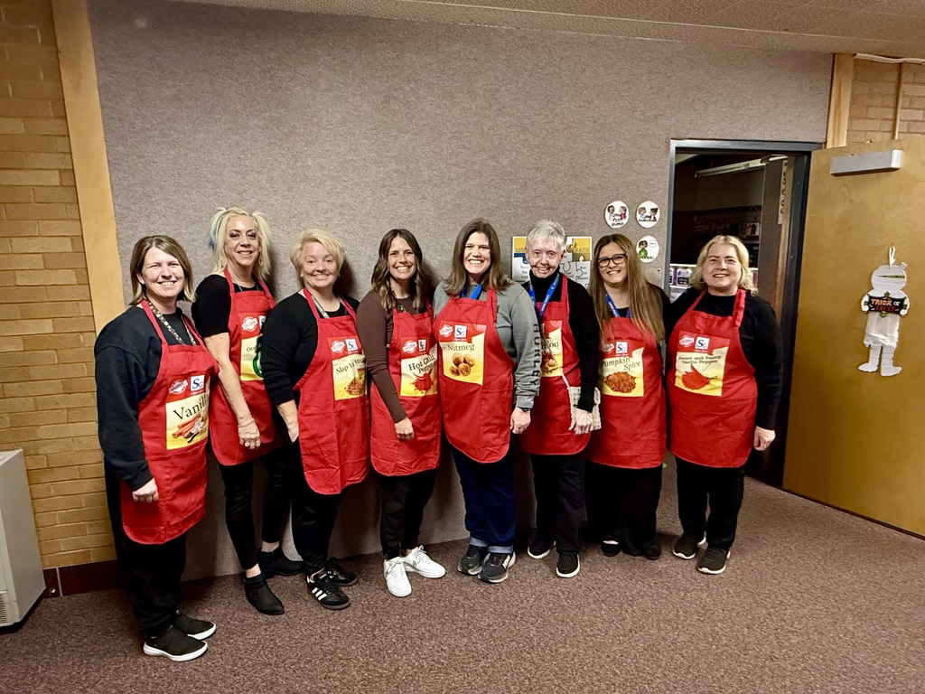 A group of approximately 6-8 elementary school staff members (women) dressed in matching bright red aprons, each labeled with a different spice or seasoning like Vanilla, Hot Chili Pepper, Nutmeg, and Pumpkin Spice, for a group Halloween costume. They are smiling in a hallway and classroom setting.