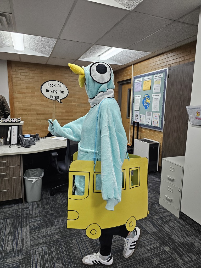 An elementary school staff member in an office setting dressed in a creative costume as "The Pigeon" from Mo Willems' children's book, wearing a large blue bird costume and a yellow school bus structure around their waist, holding a sign that reads, "Look, I'm driving the bus!!!"