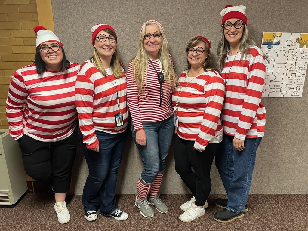 Five female teachers, all dressed as the character Waldo/Wally (red and white striped shirts, glasses, and hats), smiling and posing for a group photo in a school hallway.
