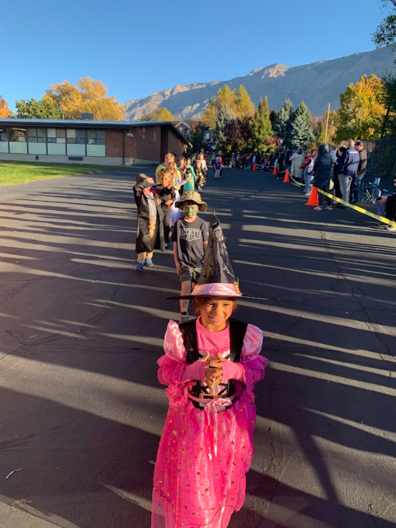 Students in costumes lined up outside on the blacktop for the parade, with a bright blue sky and mountains in the background. A girl in a bright pink princess costume and witch's hat is in the foreground.