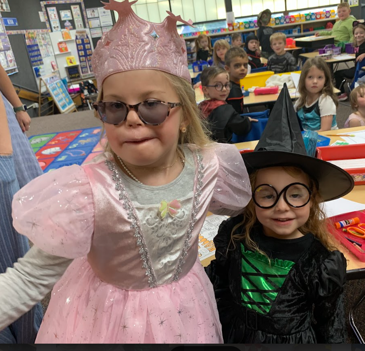 A close-up of two young girls in a classroom: one dressed as a pink princess wearing sunglasses and a crown, and the other as a witch wearing glasses.