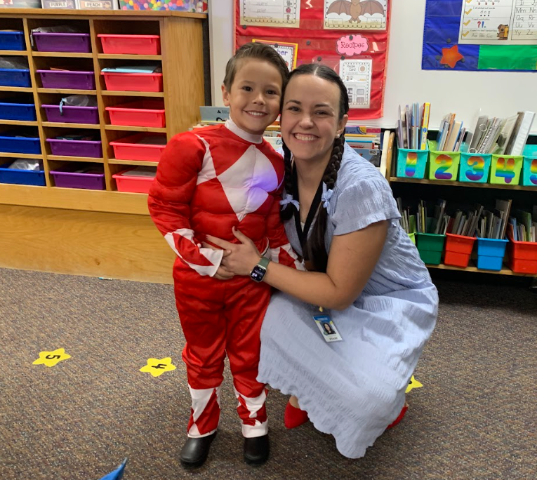 A teacher, possibly dressed as Dorothy from the Wizard of Oz, kneels next to a smiling student dressed as the Red Power Ranger in a classroom.