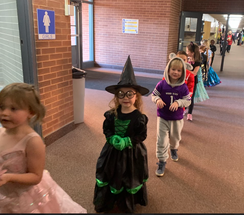 A line of young students in the school hallway during the parade. A girl in a black and green witch costume with glasses smiles directly at the camera, followed by a boy in a purple and grey mouse costume.