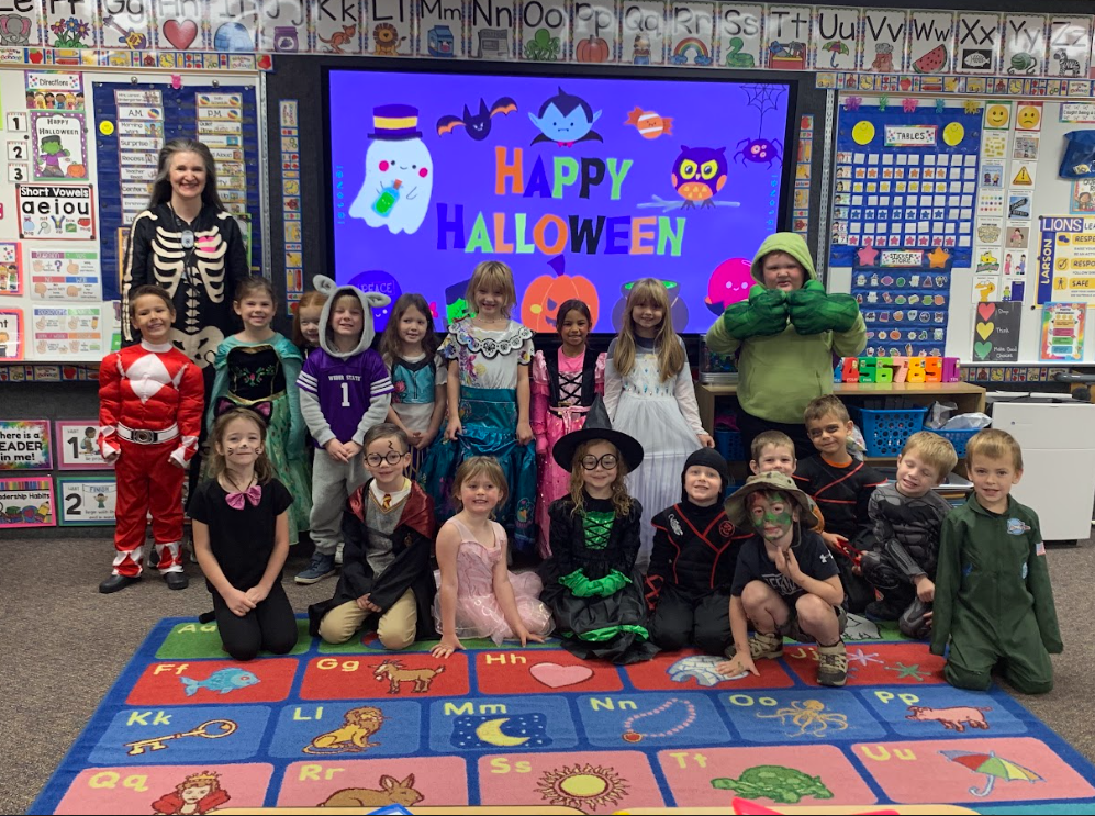 A large class photo of students in various costumes (including a Power Ranger, a mermaid, and the Hulk) posing with their teacher, who is dressed as a skeleton, in front of a smartboard with a "Happy Halloween" graphic.