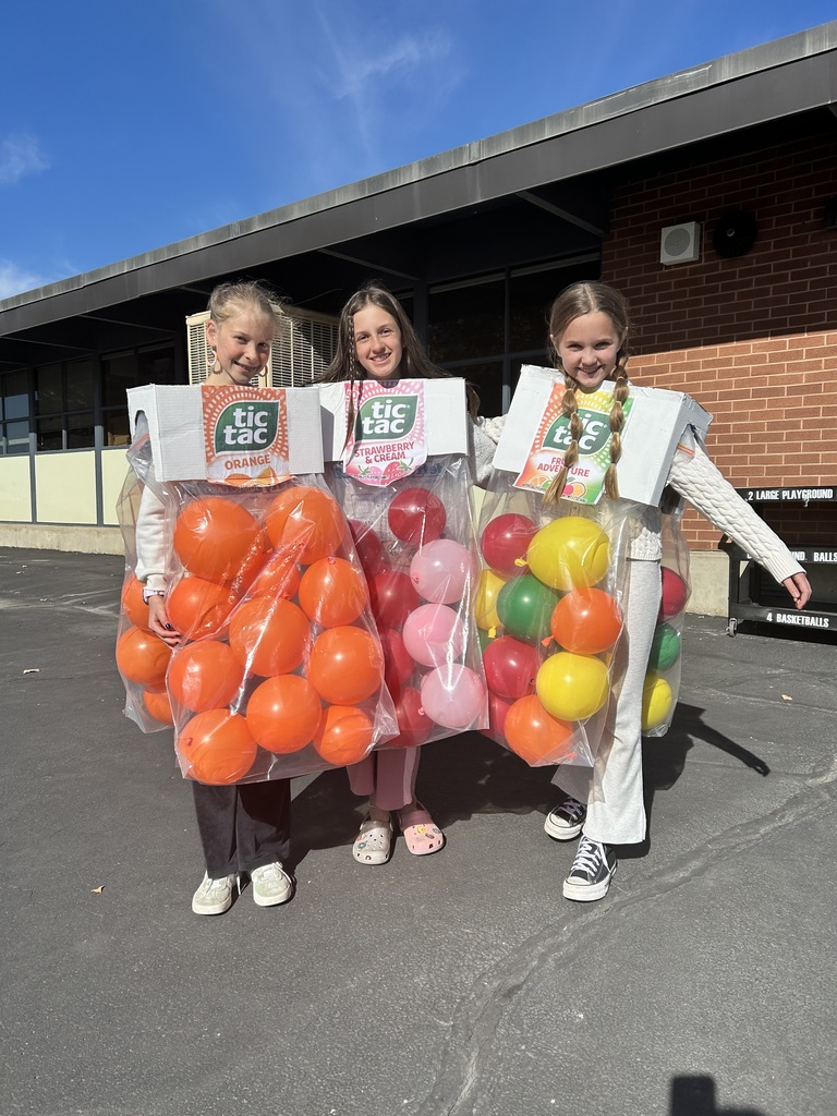 A group of three girls dressed in creative "Tic Tac" costumes (large bags filled with balloons) standing outside the school on a sunny day.