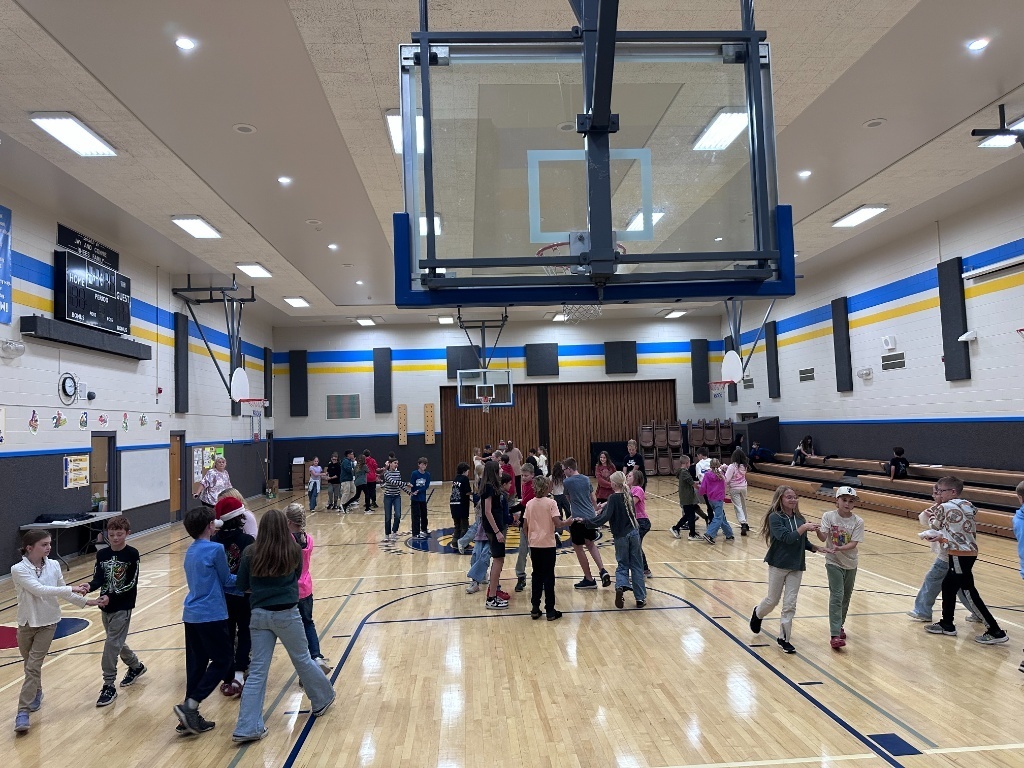 A wide shot of a brightly lit elementary school gymnasium with wooden floors. Many children, dressed in casual clothes, are moving around and engaging in a group activity on the basketball court. Basketball hoops are visible at either end.