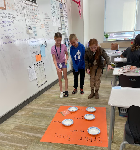 Three elementary school children, two in costume, play a "Spider Toss" game on the classroom floor, which involves tossing items into bowls placed on an orange mat with point values of "5," "10," and "50 points" written on it.