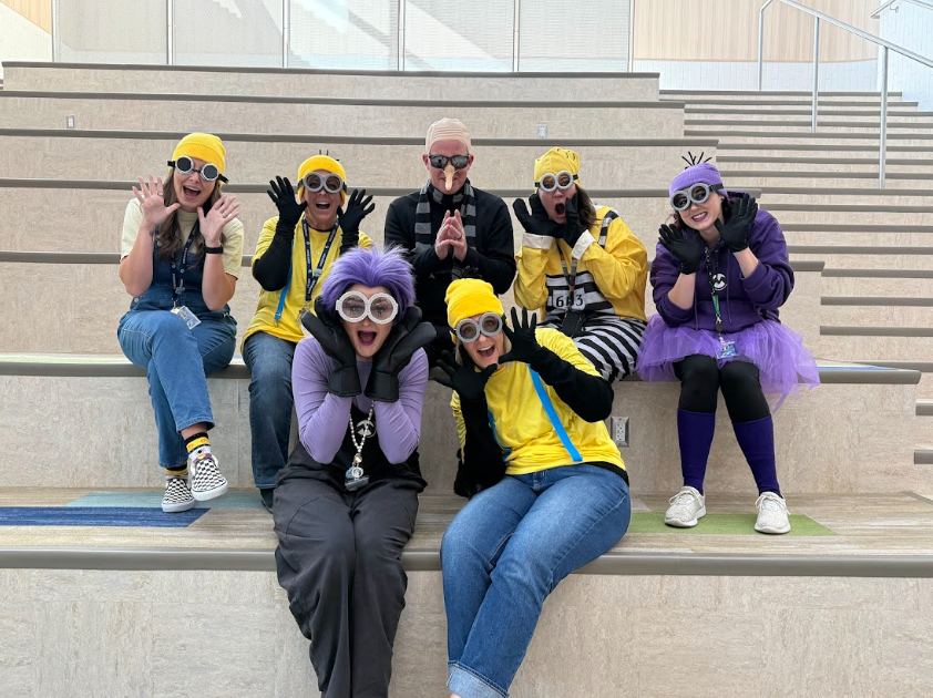 A group of six female staff members and Mr. Skeen posing on a wide staircase. All are wearing Minion-themed costumes, including yellow shirts, goggles, and yellow/purple wigs or hats. Mr. Skeen is dressed as Gru.