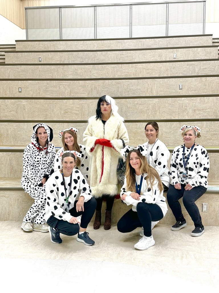 A group of seven female teachers on a set of wide, light-colored stairs. One teacher in the center is dressed as Cruella de Vil, and the six surrounding teachers are wearing spotted dalmatian-themed tops and costumes.