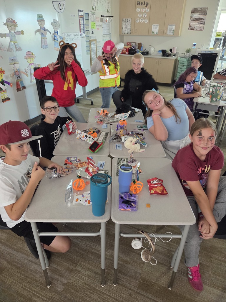 A classroom of elementary school students, many in costume (including a Minion, a construction worker, a furry creature, and Alvin from Alvin and the Chipmunks), sitting around tables covered in candy and Halloween-themed items.