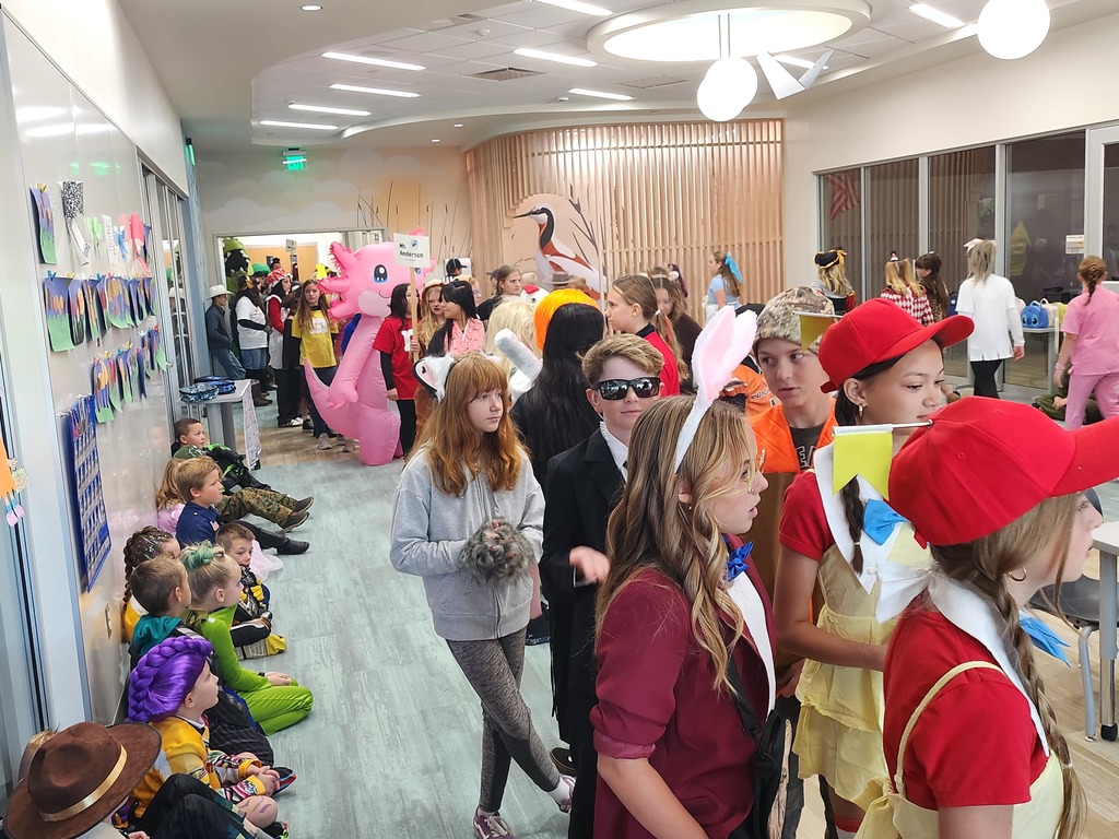 Students of various ages, many in Halloween costumes, stand and sit along a school hallway, waiting. Costumes include an inflatable pink axolotl, a person in a black suit and sunglasses, a rabbit with bunny ears, and a person in a red cap.