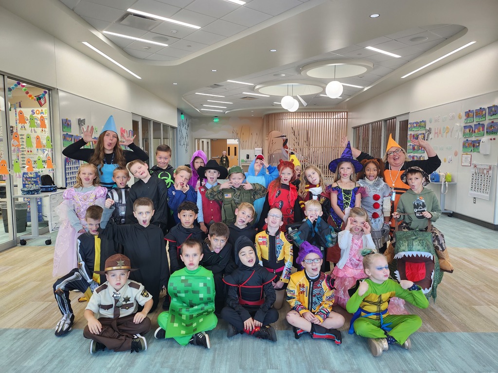 A large group of young children and a few adults, all in various Halloween costumes, posing together in a brightly lit school hallway. Costumes include ninjas, a creeper from Minecraft, a Stormtrooper, a police officer, princesses, and witches.