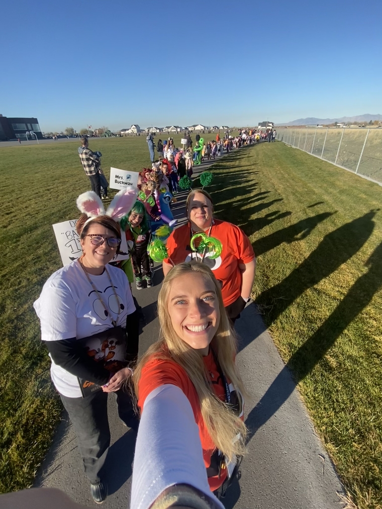 A selfie taken outdoors showing three smiling female teachers, two wearing bunny ears, standing on a sidewalk. They are part of a long line of students and teachers, many in costume, participating in a Halloween parade. A sign on the grass reads "Mrs. Buckwau."