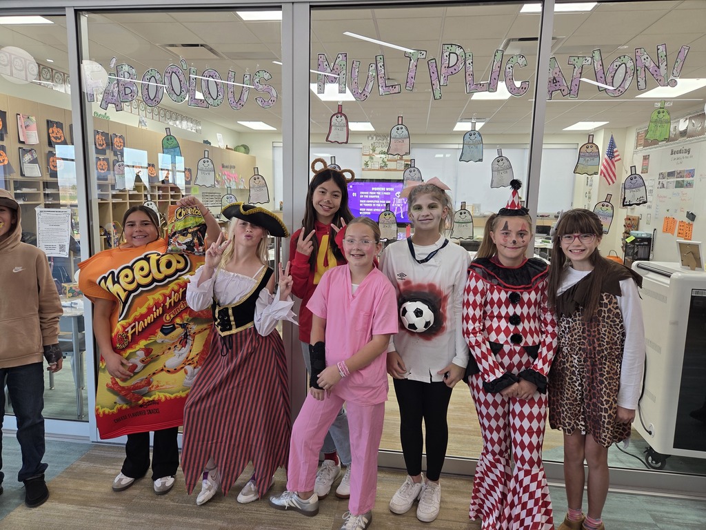 A group of seven elementary school girls in Halloween costumes, including a Cheetos bag, a pirate, a cartoon bear, a doctor/scrubs, a soccer player with pig face paint, a harlequin/clown, and a cave girl, standing in front of a glass classroom wall. The wall has cutout letters spelling "FABOOLOUS MULTIPLICATION" across the top.