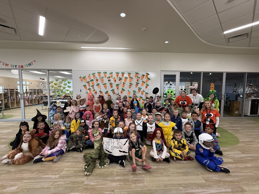 A very large group of first-grade students and their teachers, all dressed in a wide array of Halloween costumes, posing on the floor of a school room. A white sign held by a student reads: "DON'T GET CRABBY IT'S 1ST GRADE." The wall behind them is decorated with cutout carrots.
