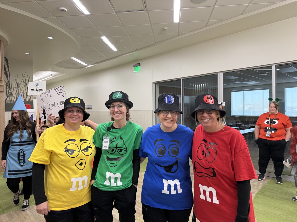 Four female lunch staff smiling, dressed in matching bucket hats and brightly colored M&M t-shirts (yellow, green, blue, and red) in a school hallway. Another teacher in an orange M&M shirt is visible behind them.