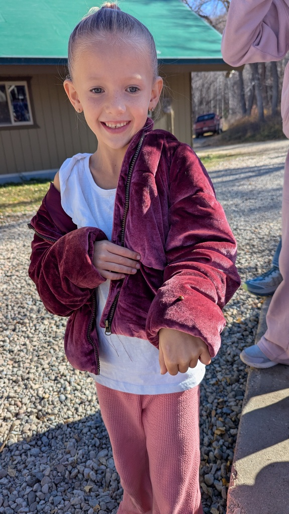 A young girl in a maroon velvet jacket and white shirt smiling brightly at the camera outdoors.