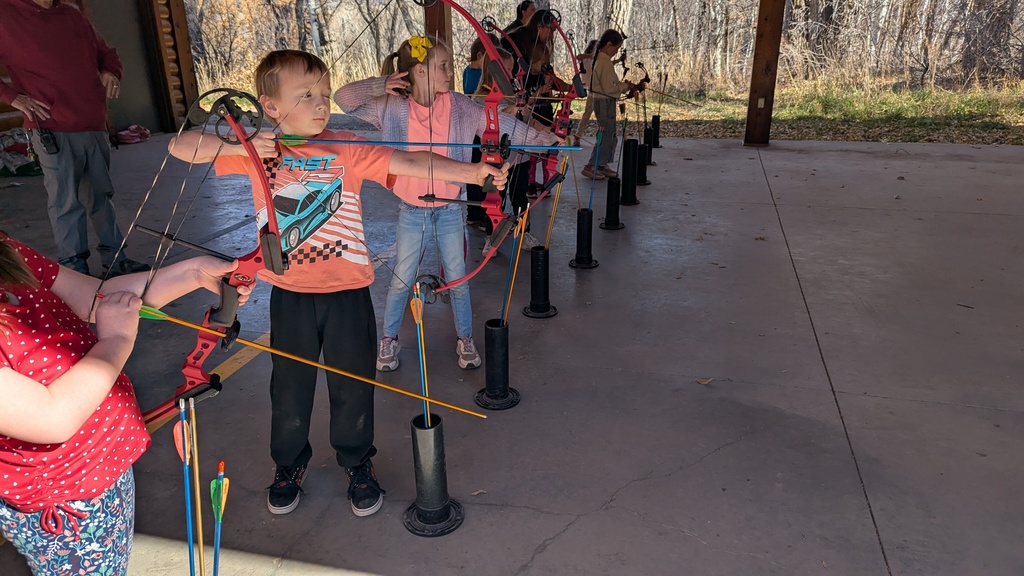 A young boy in an orange shirt with a car graphic, aiming a red and black bow and arrow at the archery range.